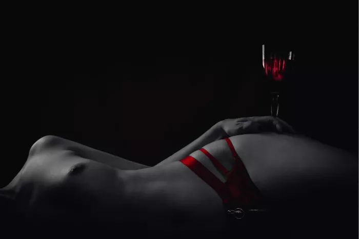 Liggende vrouw in rode lingerie met een glas rode wijn op de heup, low-key zwart-wit met rood