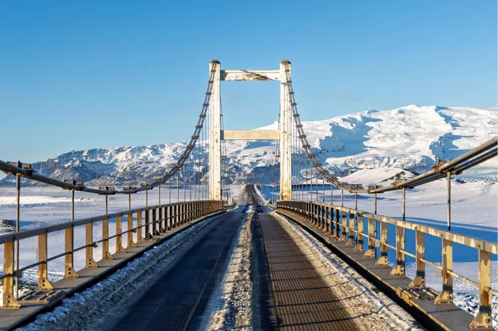Hangbrug met witte pylonen en stalen kabels over een besneeuwde vlakte, met witte bergen en een blauwe lucht
