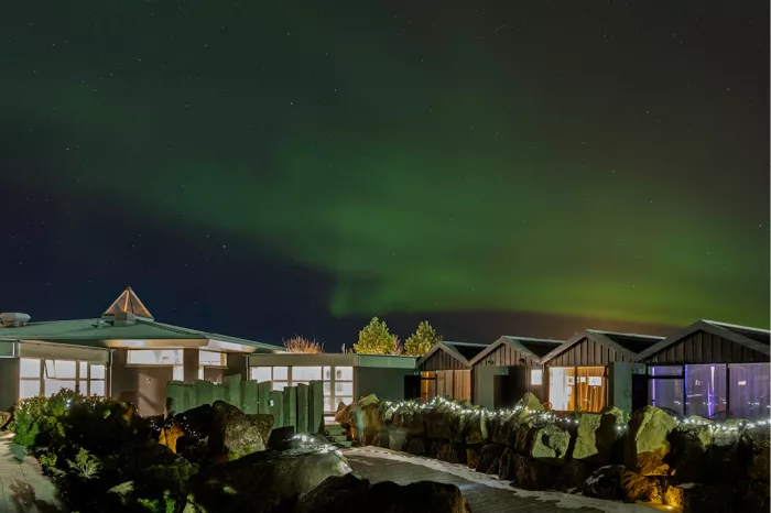 Groen noorderlicht boven lage houten huisjes met lichtsnoeren en rotsen bij nacht