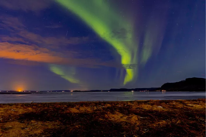 Groen noorderlicht in een boog boven het ijs, met oranje wolken en een donkere heuvelrug