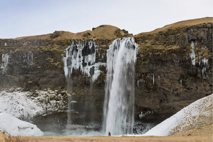 Bevroren waterval met ijspegels langs een bruine rotswand en een klein figuurtje aan de voet