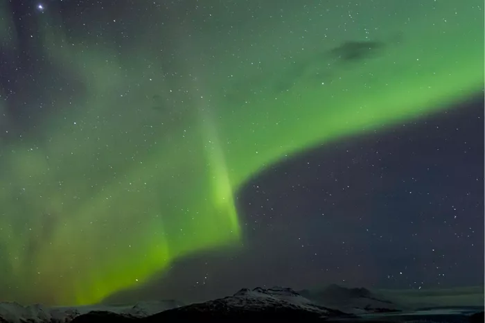 Brede groene banen noorderlicht en sterren boven een smalle rand besneeuwde bergen