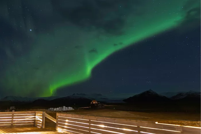 Groene boog noorderlicht boven een verlicht houten terras met bergen in het donker