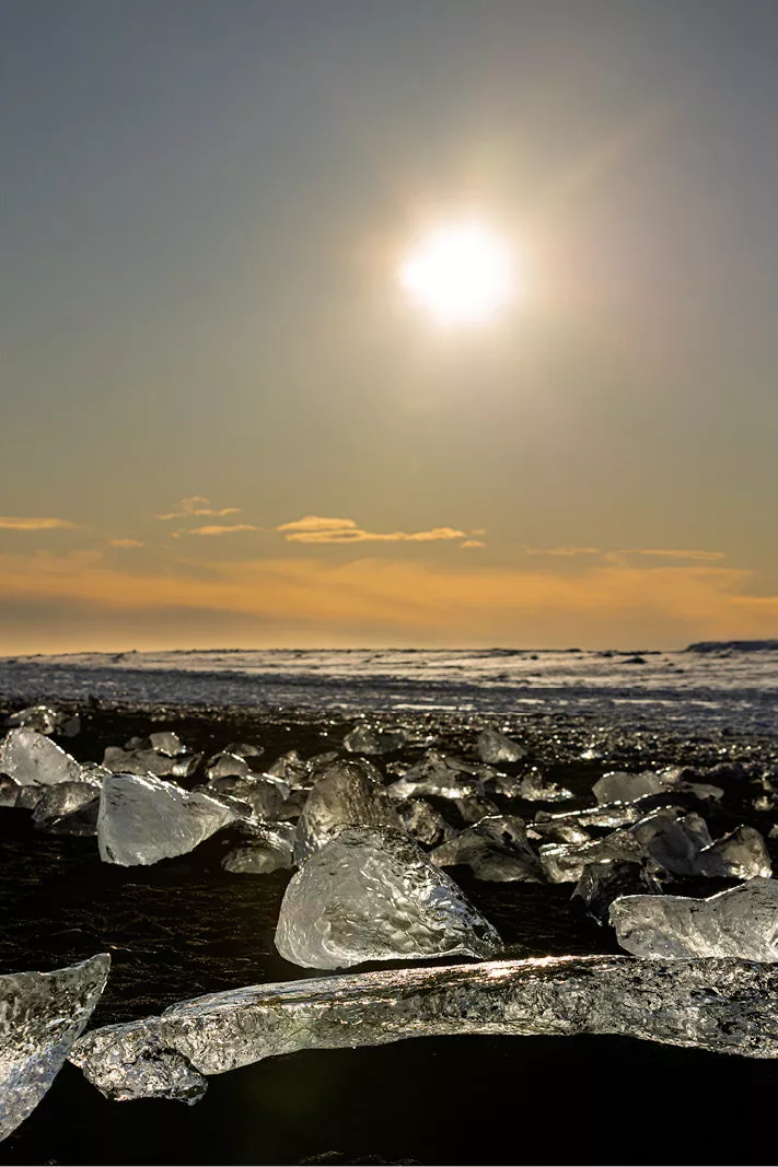 Glinsterende ijsblokken op een zwart strand in tegenlicht, met oranje wolken aan de horizon