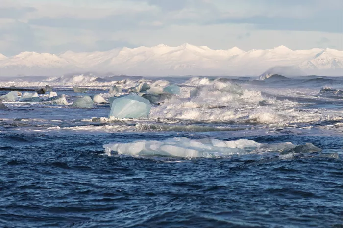 Blauwe ijsblokken in een woelige blauwe zee met besneeuwde bergtoppen erachter