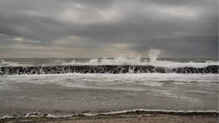 Golven die bij donker weer opspatten tegen een strekdam van zwarte stenen onder een grijze wolkenlucht