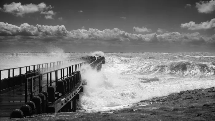 Woeste golven die tijdens een zomerstorm over een houten pier met reling slaan, in zwart-wit
