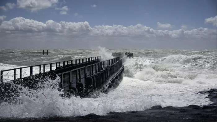 Witte golven die tegen een donkere houten pier slaan onder een blauwe lucht met stapelwolken