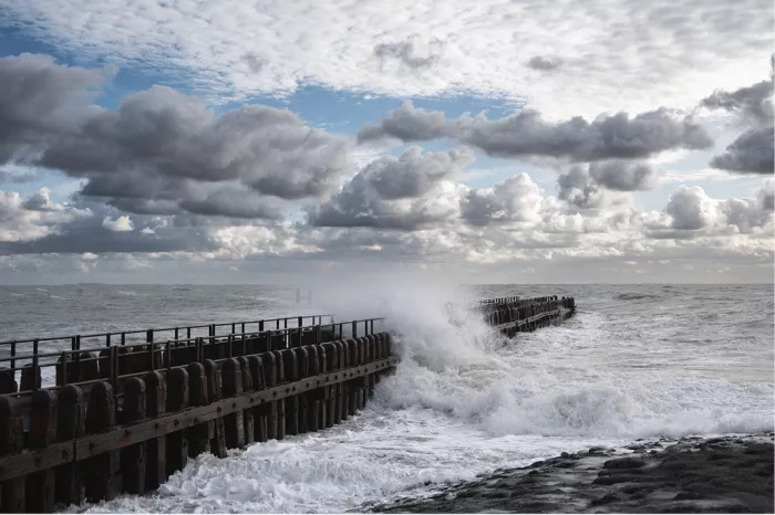 Lange houten pier met golven die hoog opspatten onder grote grijze en witte wolken