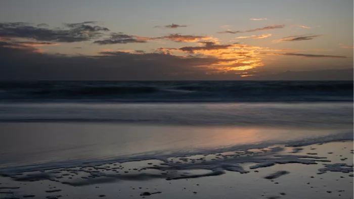 Zonsondergang achter donkere wolken boven zee met een nat strand vol plasjes in het laatste licht