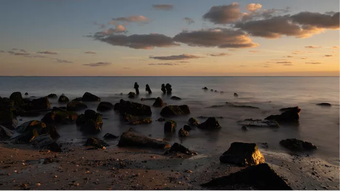 Donkere stenen en paaltjes in zee en op het strand, met warm licht van de ondergaande zon