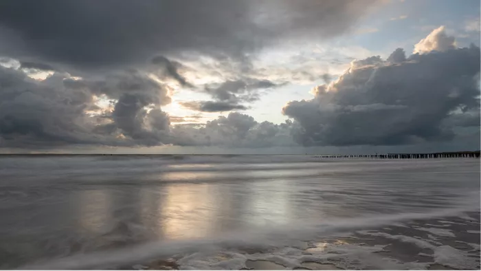 Zilverachtig licht dat in de zee spiegelt onder grijze wolken met paalhoofden aan de horizon
