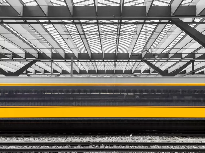 Vervaagde trein met gele strepen onder een stationsoverkapping, rest in zwart-wit