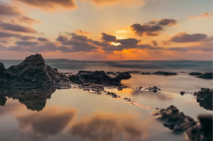 Een zonsondergang aan de kust, met de zon die ondergaat aan de horizon. De lucht is gevuld met oranje, roze en paarse tinten, die worden weerspiegeld in het water op de voorgrond. Rotsformaties zijn zichtbaar langs de kustlijn, met de golven die zachtjes over de rotsen slaan.