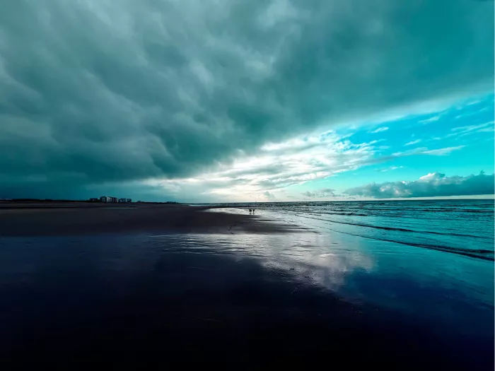 Strand met de zee aan de rechterkant. De lucht is dramatisch met donkere wolken aan de linkerkant en lichtere, helderblauwe lucht aan de rechterkant.