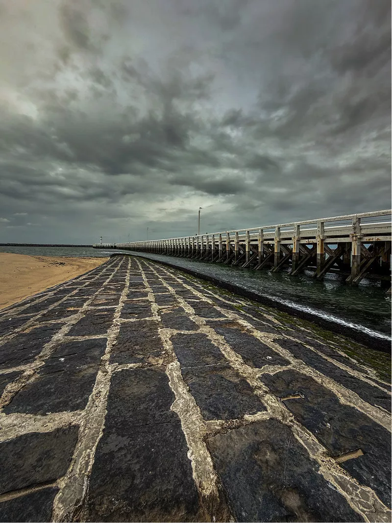 Stenen golfbreker naast een houten pier onder een grijze wolkenlucht