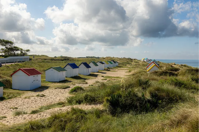 Rij witte strandhuisjes met gekleurde daken in de duinen, met helmgras en zee onder wolken