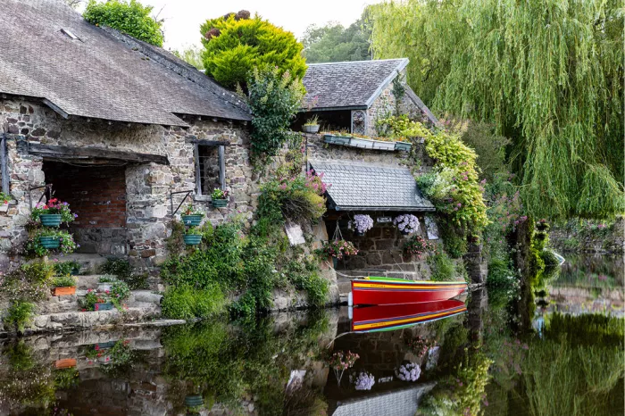 Rode gestreepte roeiboot voor een oud stenen huis met bloembakken en een treurwilg, gespiegeld in het water