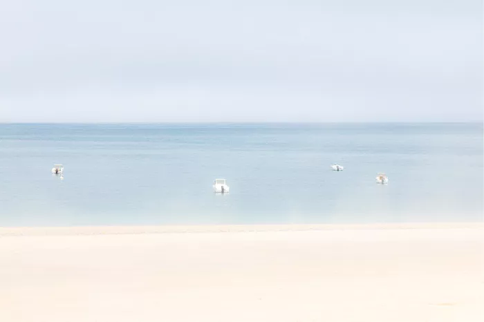 Vier kleine bootjes drijven op een kalme zee onder een zachte pastelkleurige lucht, met een lege zandstrook op de voorgrond.