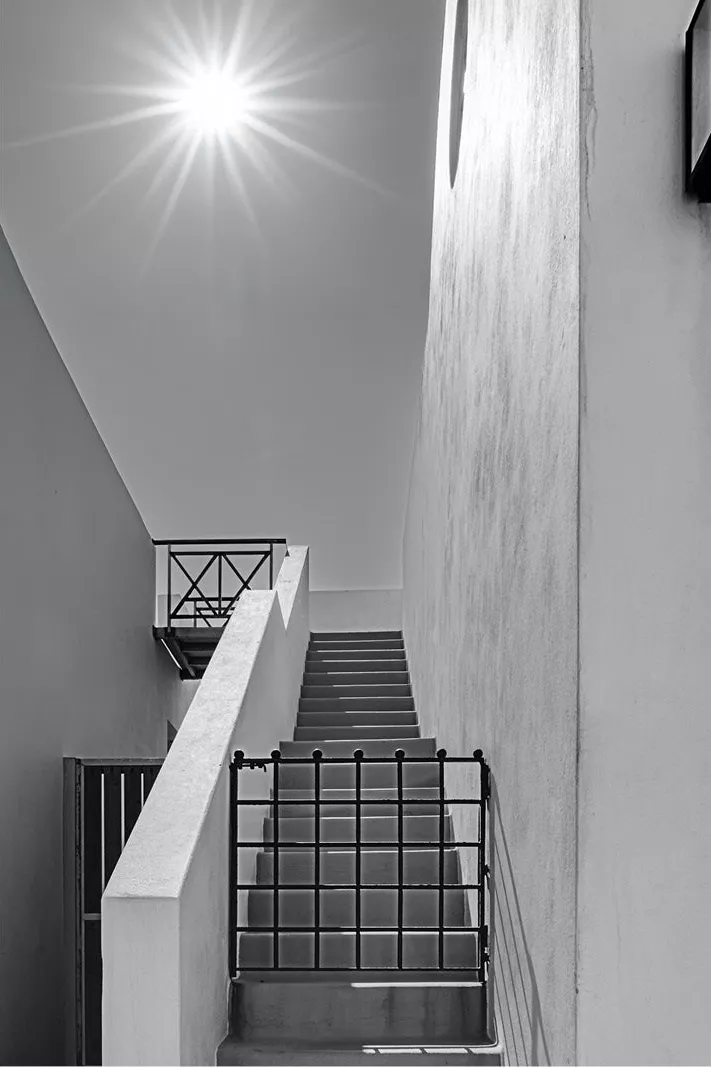 Betonnen trap tussen witte muren met een zwart hekje, en een stralende zon boven de gevel, in zwart-wit