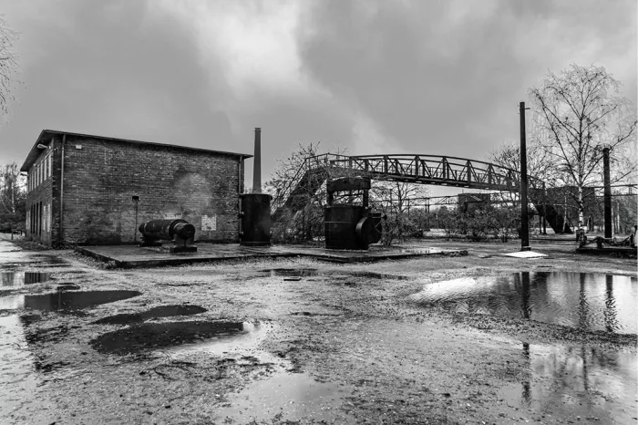 Verlaten industrieterrein met bakstenen gebouw, schoorsteen en metalen loopbrug, met plassen op de grond, in zwart-wit