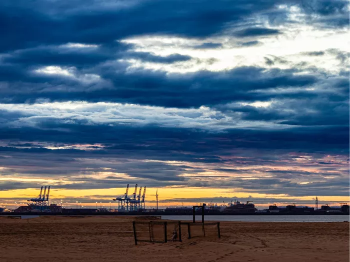Strand onder een dramatische wolkenlucht bij zonsondergang, met havenkranen aan de horizon