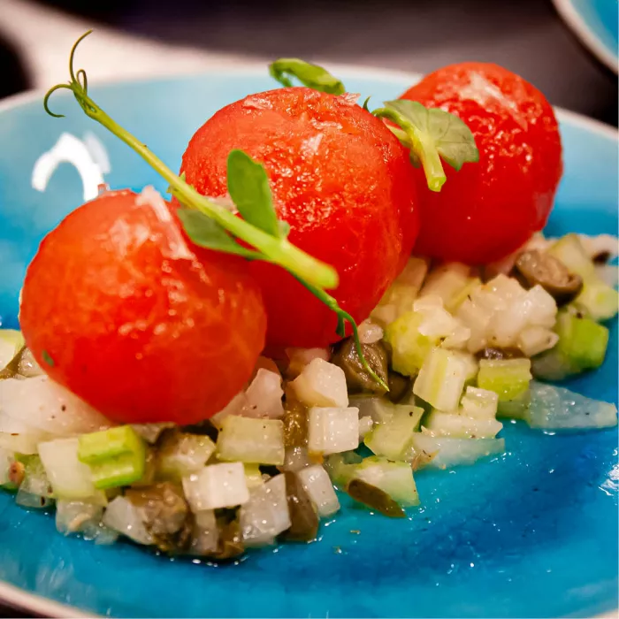 Fraai gestileerd culinair gerecht bestaande uit drie perfect gepelde, helderrode tomaatjes die elegant op een basis van fijngesneden groenten en kappertjes zijn geplaatst. 