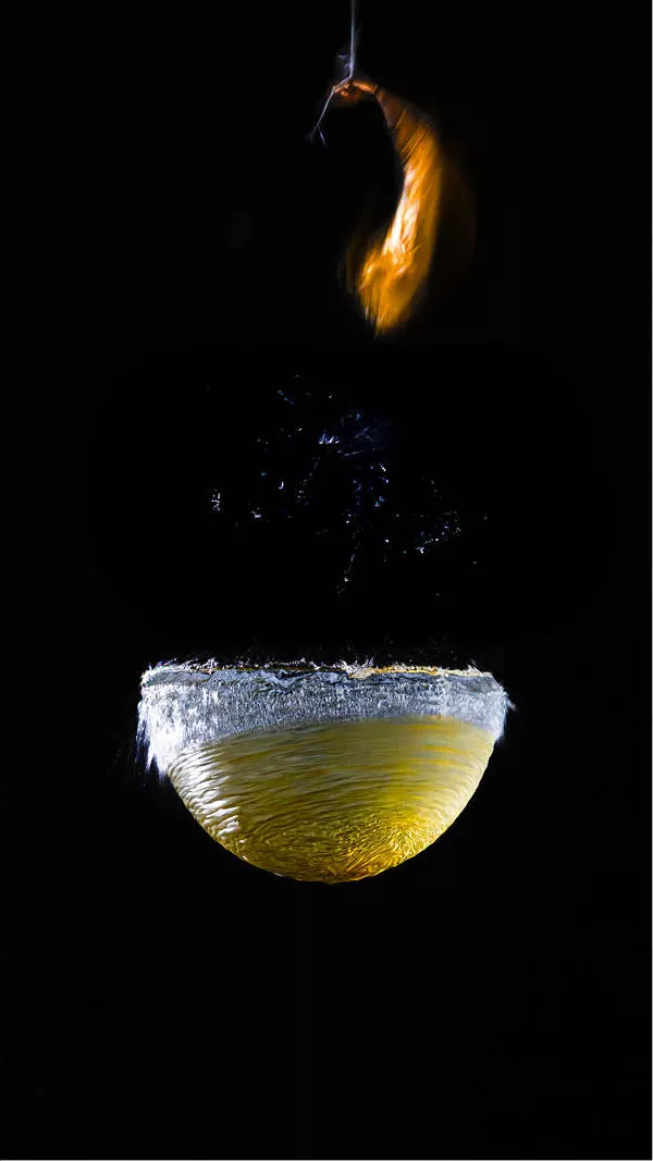 Een felgele watermassa blijft nog heel even in vorm hangen terwijl de ballon eromheen in een fractie van een seconde uit elkaar spat. De bovenste fragmenten van het ballonrubber bewegen als een lichtgevende sliert omhoog, terwijl het water onderaan een gladde, bijna glasachtige halve bol vormt. 