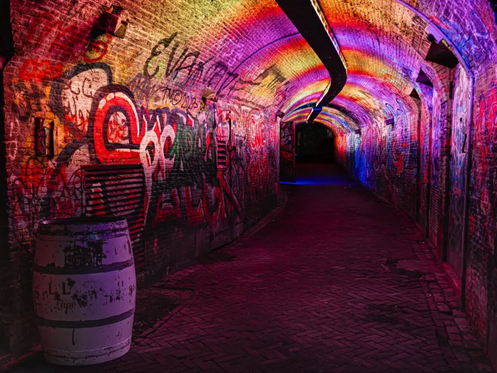 Bakstenen tunnel met gewelfd plafond baadt in regenboogkleurig licht, met graffiti op de muren, een versleten witte ton op de voorgrond links en een klinkerpad dat naar het donkere uiteinde loopt.