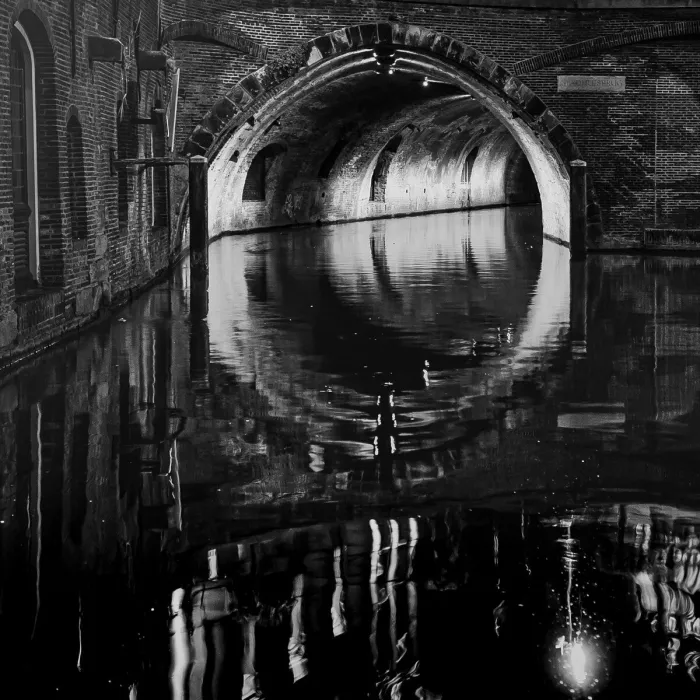 Verlichte bakstenen brugboog van de Stadhuisbrug in Utrecht, met een gewelfde tunnel boven een donkere gracht waarin de ronde boog en de lichtpunten rimpelend weerspiegeld worden in zwart-wit.