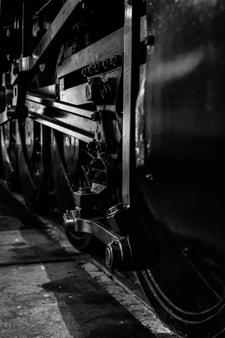 Zwart-witdetail van het onderstel van een stoomlocomotief, met glanzende drijfstangen, bouten en moeren, spaakwielen en een donkere ketelwand, waarbij schaduwen over de betonnen vloer van de loods vallen.